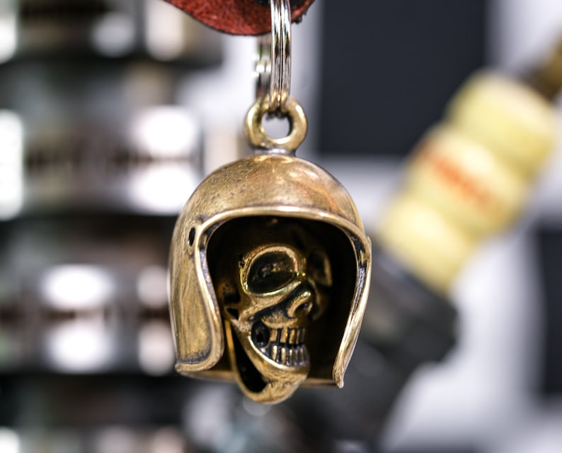 Puede incluir: Un llavero de calavera de color dorado con un dise&ntilde;o de casco. La calavera est&aacute; detallada con cuencas de los ojos, una nariz y una boca. El llavero est&aacute; unido a una correa de cuero.
