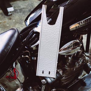 May include: A white, textured motorcycle seat cover with a unique shape, featuring two small holes at the bottom. The cover is placed on a black motorcycle with chrome accents, including the Harley Davidson logo.