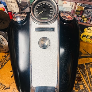 Puede incluir: Tanque de gasolina negro de motocicleta con velocímetro y detalles cromados. El velocímetro muestra MPH. El tanque tiene un panel blanco texturizado y dos tapones de combustible cromados. El fondo incluye letreros y objetos vintage.