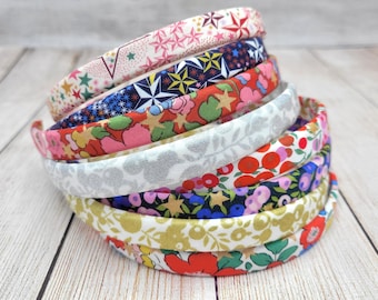 May include: A stack of eight fabric-covered headbands featuring various patterns including multicolored stars, floral prints in red, pink, and green, a silver metallic leaf design, and blue and purple berry motifs on dark and light backgrounds.