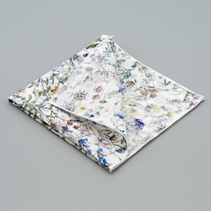 May include: A white cotton handkerchief with a floral print featuring a variety of colorful flowers and leaves. The handkerchief is folded in half and the floral print is visible on both sides.