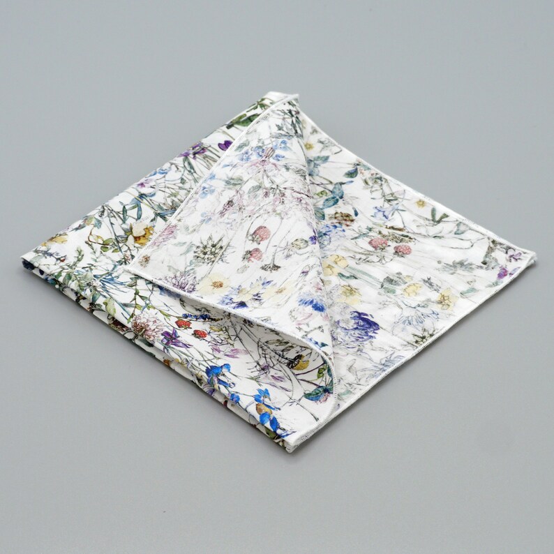 May include: A white cotton handkerchief with a floral print featuring a variety of colorful flowers and leaves. The handkerchief is folded in half and the floral print is visible on both sides.