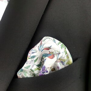 May include: A white pocket square with a floral pattern in shades of pink, green, blue, and yellow. The pocket square is folded and placed in the pocket of a black suit jacket.