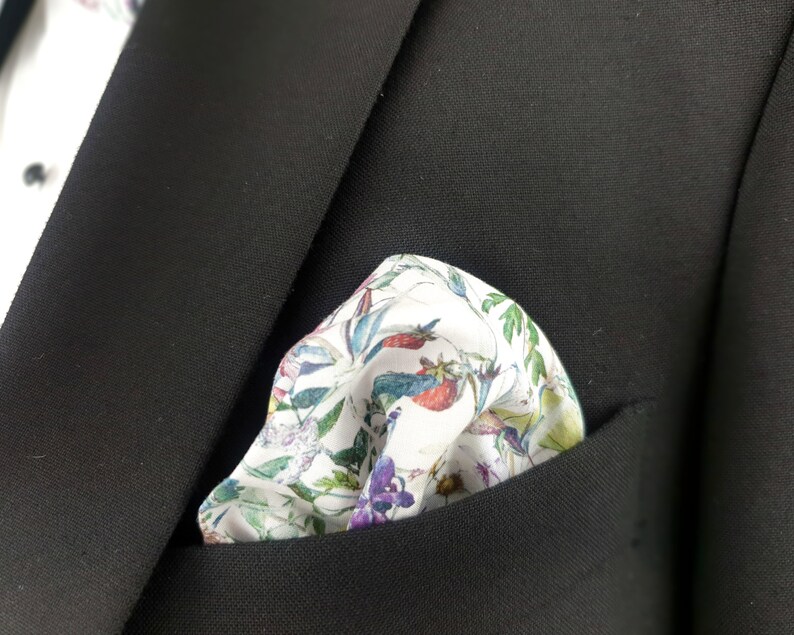 May include: A white pocket square with a floral pattern in shades of pink, green, blue, and yellow. The pocket square is folded and placed in the pocket of a black suit jacket.