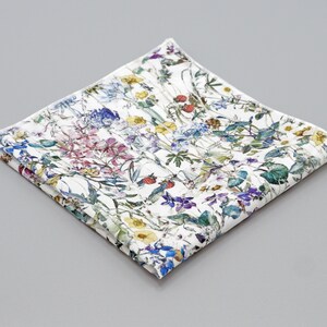 May include: A white cotton pocket square with a floral print featuring a variety of flowers and leaves in shades of blue, pink, yellow, and green.