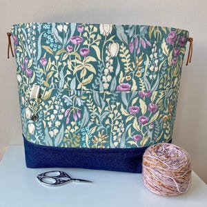 Peut inclure: Un sac à projets en tissu bleu sarcelle avec un imprimé floral et un fond en jean. Le sac a une fermeture éclair et une petite poche sur le devant. Il y a une pelote de laine et une paire de ciseaux devant le sac.