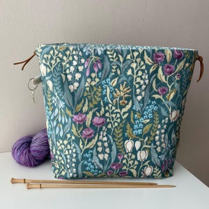 Peut inclure: Sac à ouvrage en tissu bleu sarcelle avec un motif floral composé de fleurs blanches, roses et jaunes. Le sac a des cordons de serrage en cuir marron et est posé sur une surface blanche avec des aiguilles à tricoter en bois.