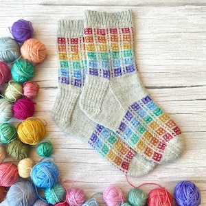 Pode incluir: Um par de meias cinzentas de malha com um padrão xadrez arco-íris no pé e na perna. As meias são rodeadas por novelos de lã coloridos em tons de roxo, laranja, verde, amarelo e azul, numa superfície de madeira.