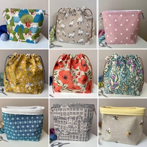 May include: A collection of nine fabric drawstring project bags featuring various patterns including mountain landscapes, llamas, polka dots, floral prints, geometric shapes, architectural building outlines, and bees on a light background.