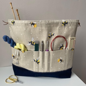 May include: A beige canvas knitting project bag with a bee print and white polka dots. The bag has multiple pockets and is filled with knitting needles, yarn, and other supplies. The bag has a navy blue bottom.