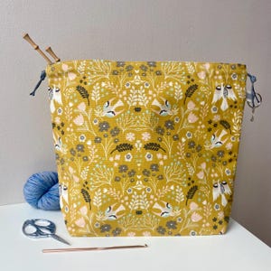 May include: Mustard yellow drawstring project bag with a floral and bird pattern. The bag has a blue drawstring closure and wooden knitting needles are inserted in the top. A ball of blue yarn, scissors, and a crochet hook are next to the bag.