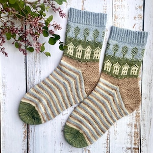 May include: A pair of knitted socks with a landscape design. The socks feature a blue cuff, a green and brown landscape with houses and trees, and striped patterns in shades of brown, blue, and cream. The heels and toes are green.
