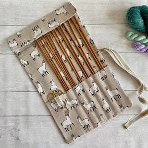 May include: A beige fabric roll-up case with a llama print and a set of wooden knitting needles. The case has a white ribbon tie and a small pair of gold scissors.