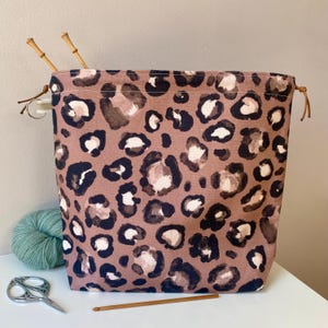 Peut inclure: Sac à fil carré à cordon de serrage avec un motif léopard dans les tons marron, noir et blanc. Le sac contient des aiguilles à tricoter en bambou. Une pelote de fil turquoise, des ciseaux et un crochet sont à côté du sac.
