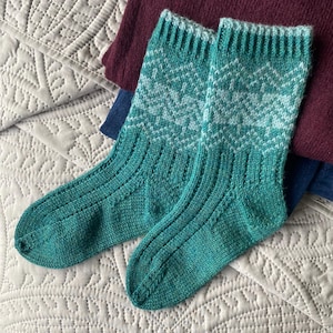 May include: A pair of teal and white patterned knit socks. The socks have a ribbed design on the foot and a chevron pattern on the leg. The socks are folded on a light gray quilted surface with a burgundy and blue fabric in the background.