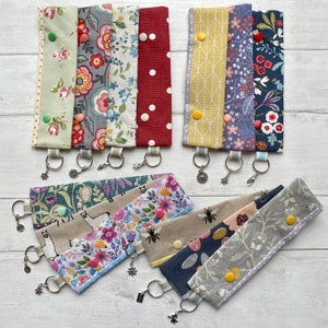 May include: A set of 10 colourful fabric keychains with different patterns. Each keychain has a silver metal ring and a silver charm. The keychains are arranged in two rows on a white surface.