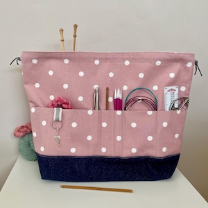May include: A pink polka dot drawstring bag with a blue denim bottom. The bag has multiple pockets and is filled with knitting needles, yarn, and other supplies.