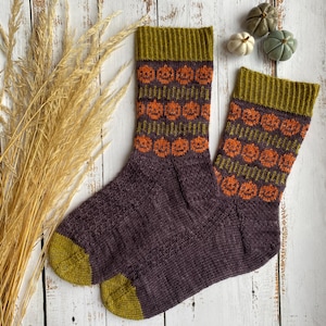 Peut inclure: Une paire de chaussettes tricotées sur le thème d'Halloween. Les chaussettes sont principalement violet foncé avec des motifs de citrouilles orange et des accents vert olive sur les poignets, les talons et les orteils. Elles sont posées sur une surface en bois clair.