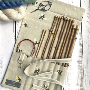 Op de afbeelding: Een beige breinaaldenrol met een bijenpatroon, met verschillende houten breinaalden, een rondbreinaald en een schaar. De rol is vastgezet met een beige band. Garen in blauwe en crèmekleurige tinten is zichtbaar.