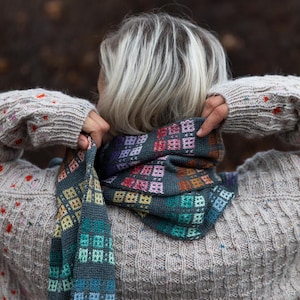 Puede incluir: Bufanda tejida a mano con un patrón geométrico en tonos turquesa, morado, naranja y amarillo. La bufanda la sostiene una persona que lleva un suéter gris. El diseño de la bufanda presenta un patrón cuadrado repetitivo.