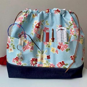May include: A light blue drawstring bag with a floral pattern in pink, red, and yellow, featuring a dark blue denim base and brown leather cords. The exterior pocket holds knitting needles, a crochet hook, and a white plastic needle gauge.