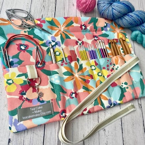 Può includere: Un astuccio per ferri da maglia con stampa floreale con vari strumenti per lavorare a maglia. La custodia è aperta, rivelando aghi colorati, uncinetti e ferri circolari. Sono visibili anche un paio di forbici e del filo. La custodia ha una cinghia e il testo "Tea Cake + Make teacakemake.Etsy.com".
