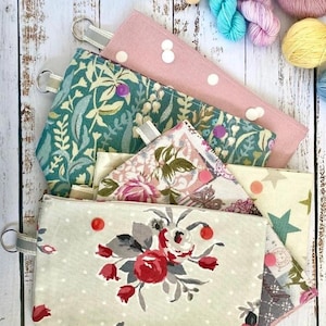 May include: Several fabric pouches with diverse patterns, such as floral, polka dots, and botanical prints. Each pouch features a metal ring and a fabric loop. The colour scheme includes pink, green, and cream. Yarn skeins in blue, pink, and yellow are visible in the background.