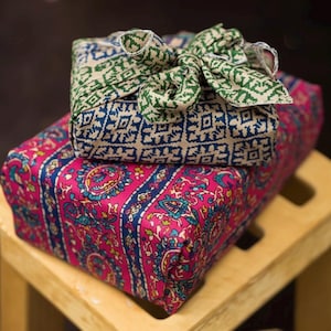 Puede incluir: Dos regalos envueltos en tela, uno encima del otro. El regalo inferior está envuelto en una tela rosa vibrante con un estampado floral y detalles azules. El regalo superior está envuelto en una tela estampada azul y beige y atado con un lazo verde.