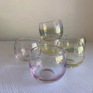 May include: Five clear, stemless drinking glasses with a rounded shape. The glasses have an iridescent sheen, reflecting pink, yellow, and green. They are arranged on a white surface, creating a subtle play of light.