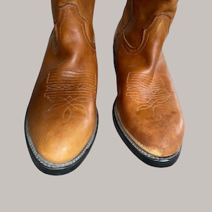 Könnte beinhalten: Ein Paar hellbraune Cowboy-Stiefel aus Leder. Die Stiefel haben dekorative Nähte auf dem Vorderblatt und dem Schaft. Die Sohlen sind schwarz mit silberner Umrandung. Das Leder weist Gebrauchsspuren auf.