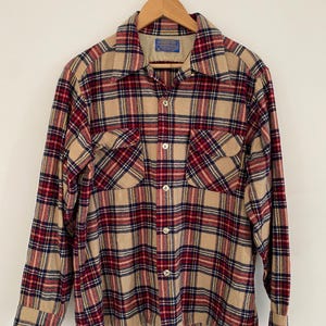 May include: A beige, red, and navy checked shirt with two breast pockets. The shirt is suspended from a wooden hanger. The shirt has a classic, vintage design.