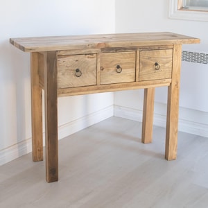May include: A wooden console table with three drawers and a natural wood finish. The table has a simple design with black metal drawer pulls.