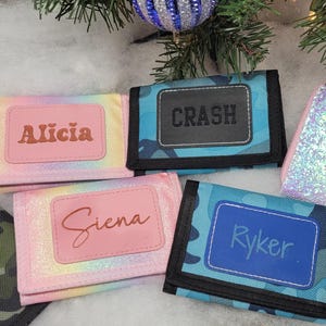 May include: A collection of personalized wallets and a zippered purse. The wallets feature names like "Alicia", "Crash", "Siena", and "Ryker" on pink or blue nameplates. The purse is iridescent with the name "Brooklyn".