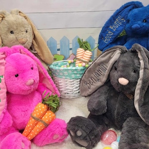 May include: Four plush bunny rabbits in beige, pink, blue, and gray. Each bunny has long floppy ears, with embroidered names or phrases. The pink bunny holds a bundle of carrot toys. A white basket with Easter eggs is in the background.