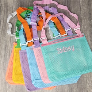May include: Six colorful mesh bags with adjustable straps and zipper closures. The bags are lined up in a row, with the straps overlapping. The bag in the front is turquoise and has the name "Sidney" embroidered on it in pink.
