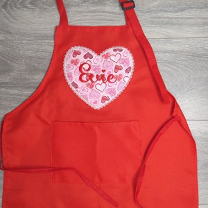 May include: Red apron with a large pink heart embroidered with the name "Evie" and a pocket. The heart is filled with smaller pink hearts.