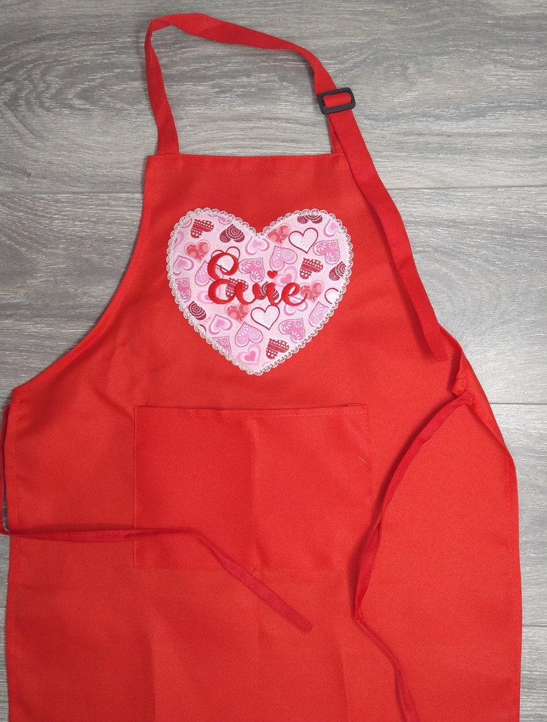 May include: Red apron with a large pink heart embroidered with the name "Evie" and a pocket. The heart is filled with smaller pink hearts.