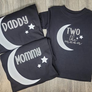 May include: Black t-shirts with silver text and moon graphics. One shirt says "Daddy", another says "Mommy", and the third says "Two the moon". Each shirt has a crescent moon and star design.