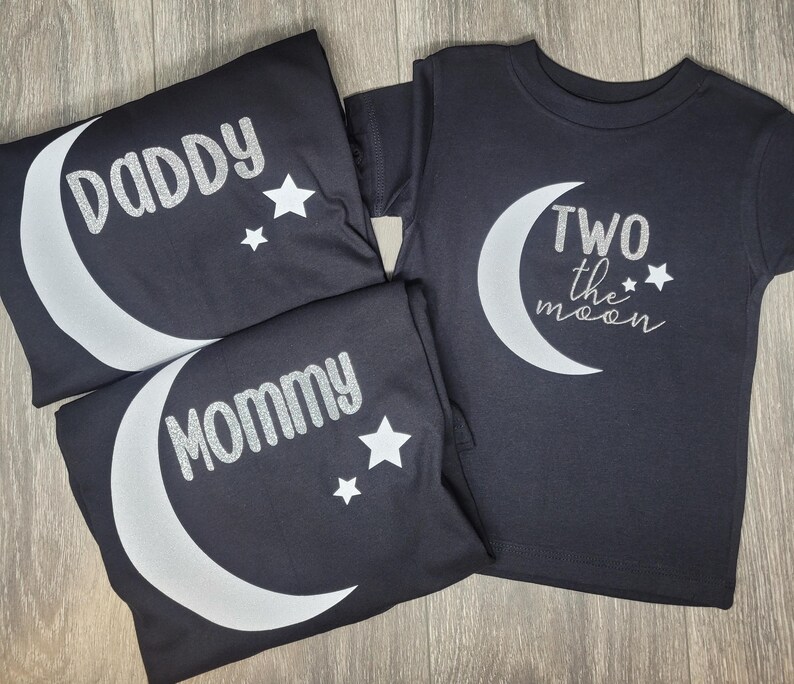 May include: Black t-shirts with silver text and moon graphics. One shirt says "Daddy", another says "Mommy", and the third says "Two the moon". Each shirt has a crescent moon and star design.