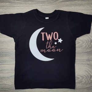 May include: Black short-sleeved t-shirt with a white crescent moon and the text "TWO the moon" in rose gold glitter. Two small white stars are next to the text. The shirt is designed for a child's second birthday.