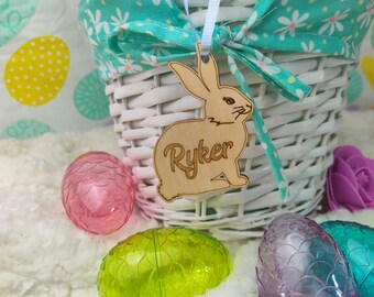 May include: A white wicker Easter basket with a teal floral fabric lining and bow. A wooden bunny tag with the name "Ryker" hangs from the basket. Colorful plastic Easter eggs in pink, green, and purple are scattered around the basket.