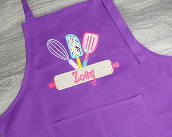 Könnte beinhalten: Lilafarbene Kinderschürze mit einer Tasche und verstellbaren Nackenriemen. Die Schürze hat ein gesticktes Design mit einem Nudelholz, einem Schneebesen und einem Spatel mit dem Namen "Zoey" auf dem Nudelholz.