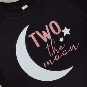 May include: Black t-shirt with a white crescent moon and the phrase "TWO the moon" in glittery rose gold. Two white stars are also featured on the shirt. The shirt is made of a soft material.