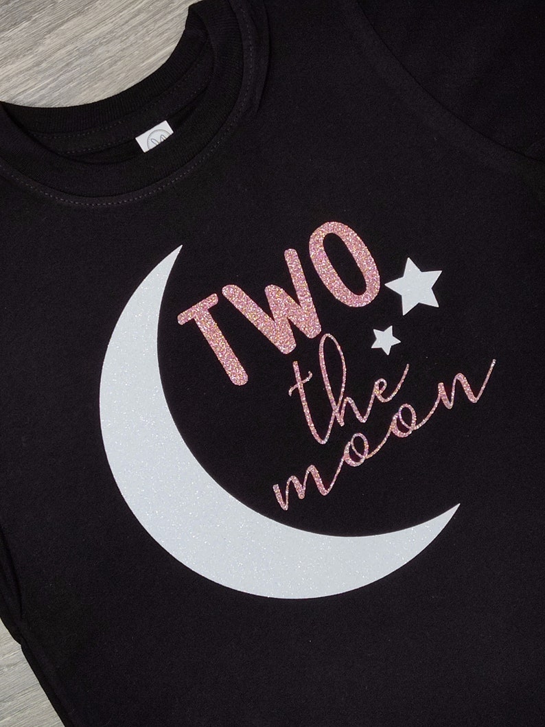 May include: Black t-shirt with a white crescent moon and the phrase "TWO the moon" in glittery rose gold. Two white stars are also featured on the shirt. The shirt is made of a soft material.