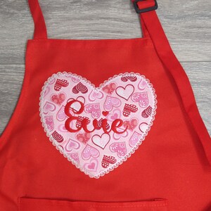 May include: Red apron with a large heart-shaped pocket featuring a white lace trim and a pink and red heart pattern. The name "Evie" is embroidered in red on the heart.