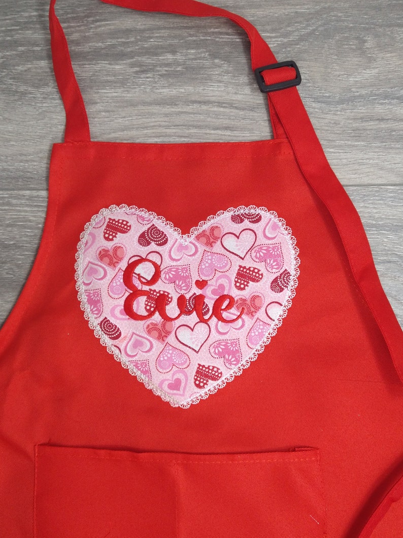May include: Red apron with a large heart-shaped pocket featuring a white lace trim and a pink and red heart pattern. The name "Evie" is embroidered in red on the heart.