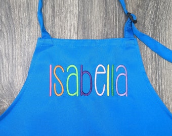 Könnte beinhalten: Eine blaue Kinderschürze mit einer großen Tasche und verstellbaren Trägern. Der Name "Isabella" ist in Regenbogenfarben auf der Vorderseite gestickt.