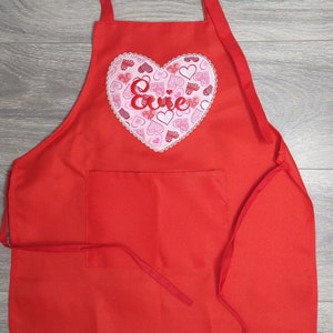May include: Red apron with a large heart-shaped pocket featuring a pink and white heart design with the name "Evie" embroidered in red.