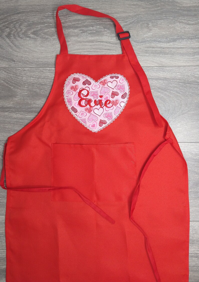 May include: Red apron with a large heart-shaped pocket featuring a pink and white heart design with the name "Evie" embroidered in red.