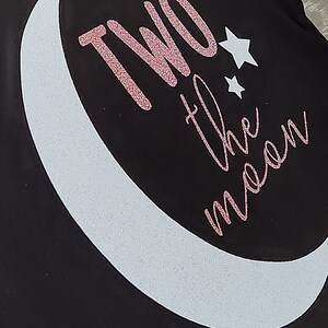 May include: Black fabric with a white crescent moon and two stars. The phrase "TWO the moon" is written in pink glitter script. The design is likely for apparel or accessories.